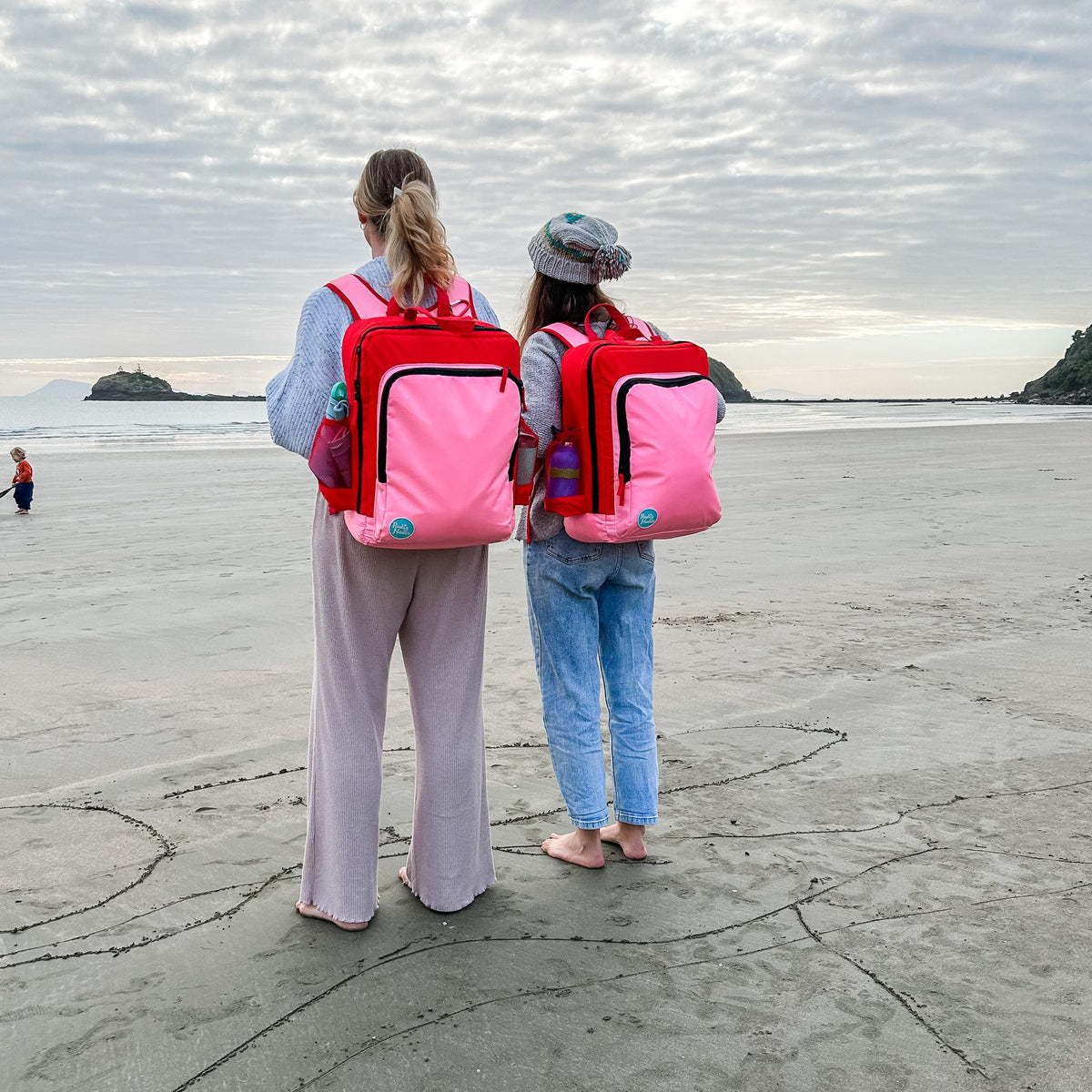 mother and child walking on the beach each wearing a blush on red adventure backpack