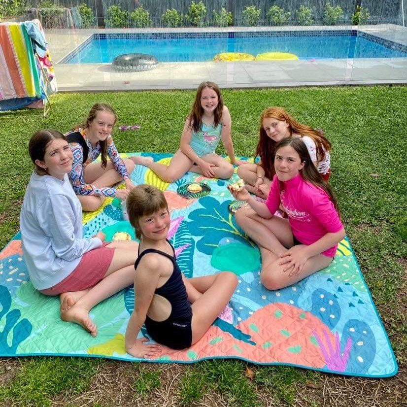 group of kids sitting on the flamingo-a-go-go waterproof picnic blanket by the pool