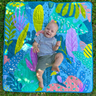 overhead view of a smiling baby lying on the flamingo-a-go-go regular waterproof playmat on the grass