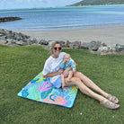 mother and baby sitting on the flamingo-a-go-go regular waterproof playmat by the beach