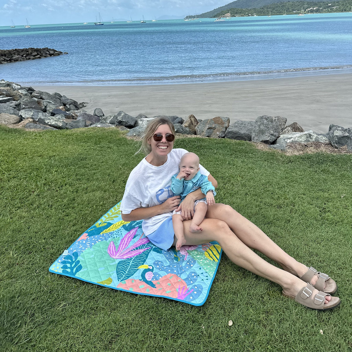 mother and baby sitting on the flamingo-a-go-go regular waterproof playmat by the beach