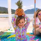 child holding a pineapple on her head sitting on the flamingo-a-go-go picnic blanket with umbrella hole at the beach