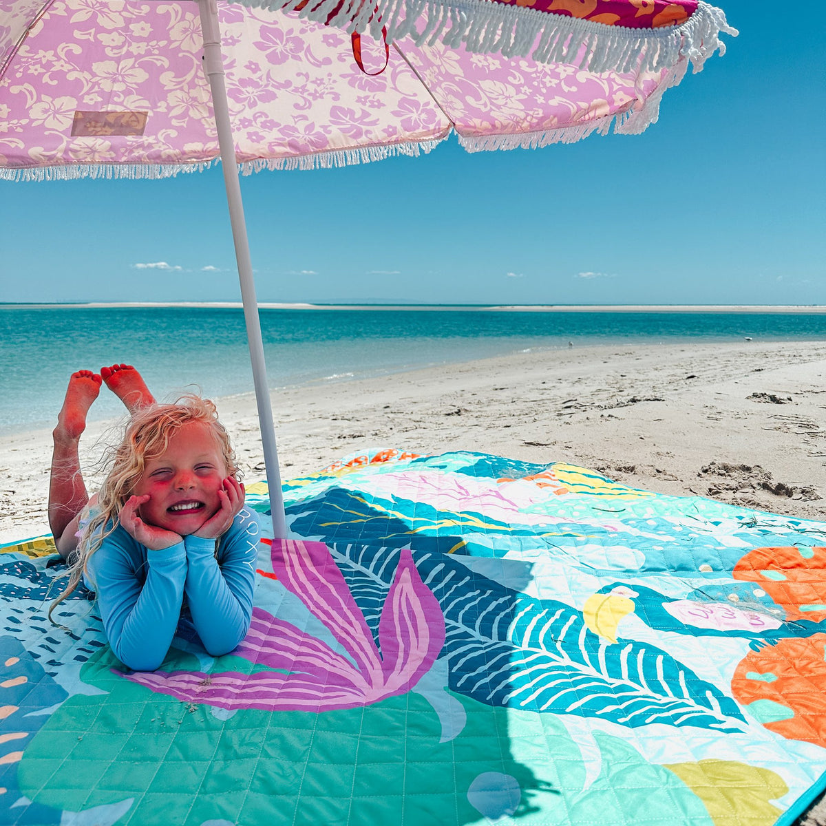 smiling child lying under a beach umbrella on the flamingo-a-go-go picnic blanket with umbrella hole