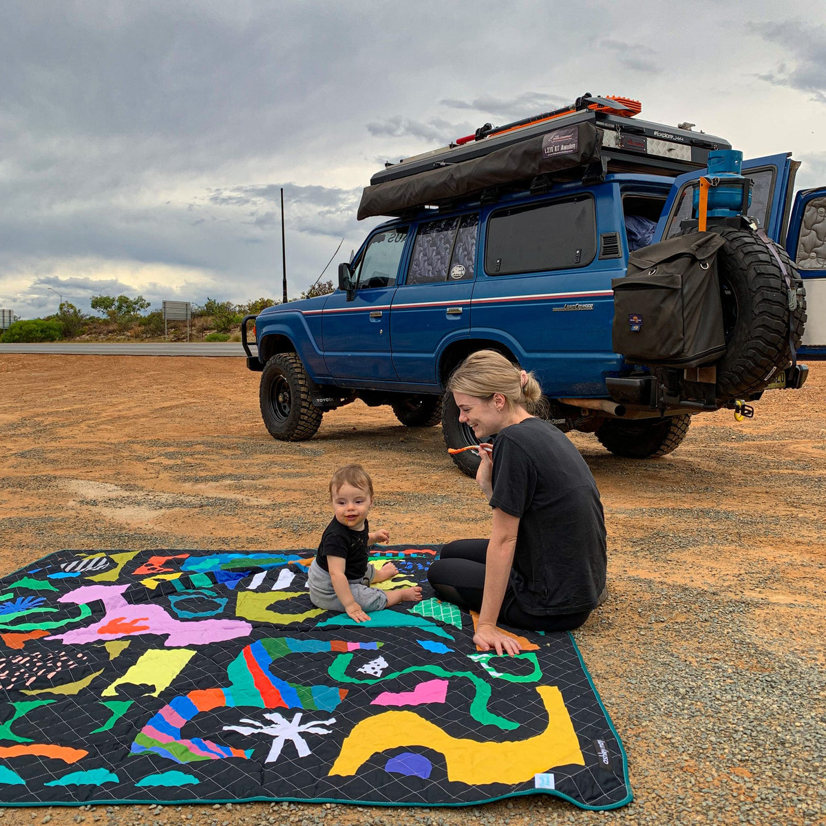 mother and baby on the rock candy waterproof picnic blanket beside a four-wheel drive on a camping trip
