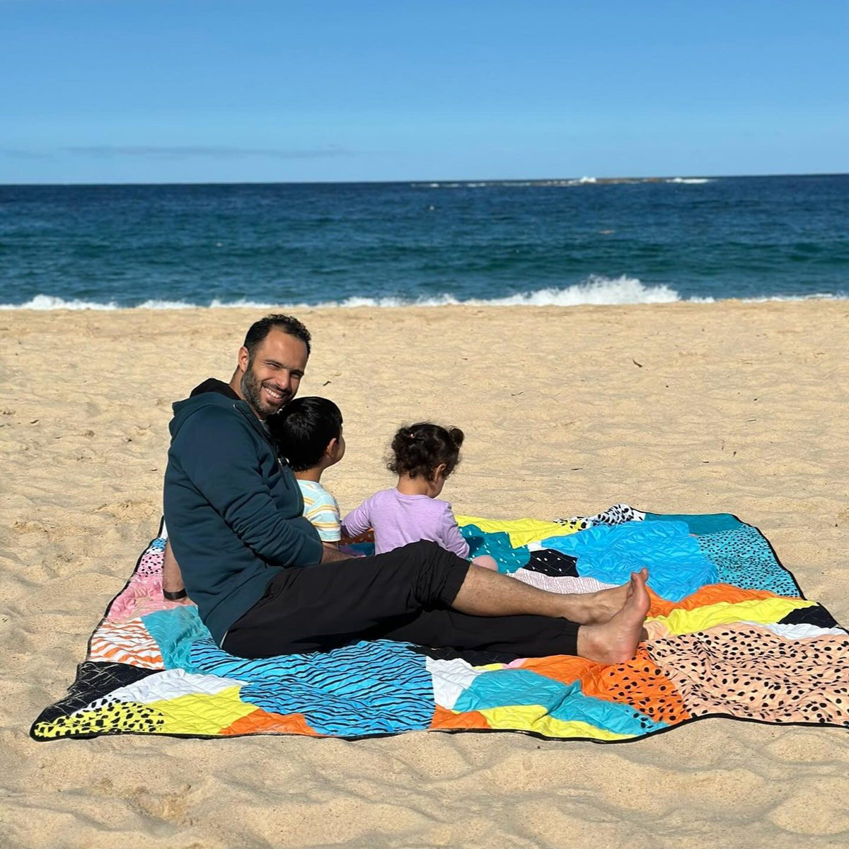 dad and two children sitting on the happy now waterproof picnic blanket on the sand